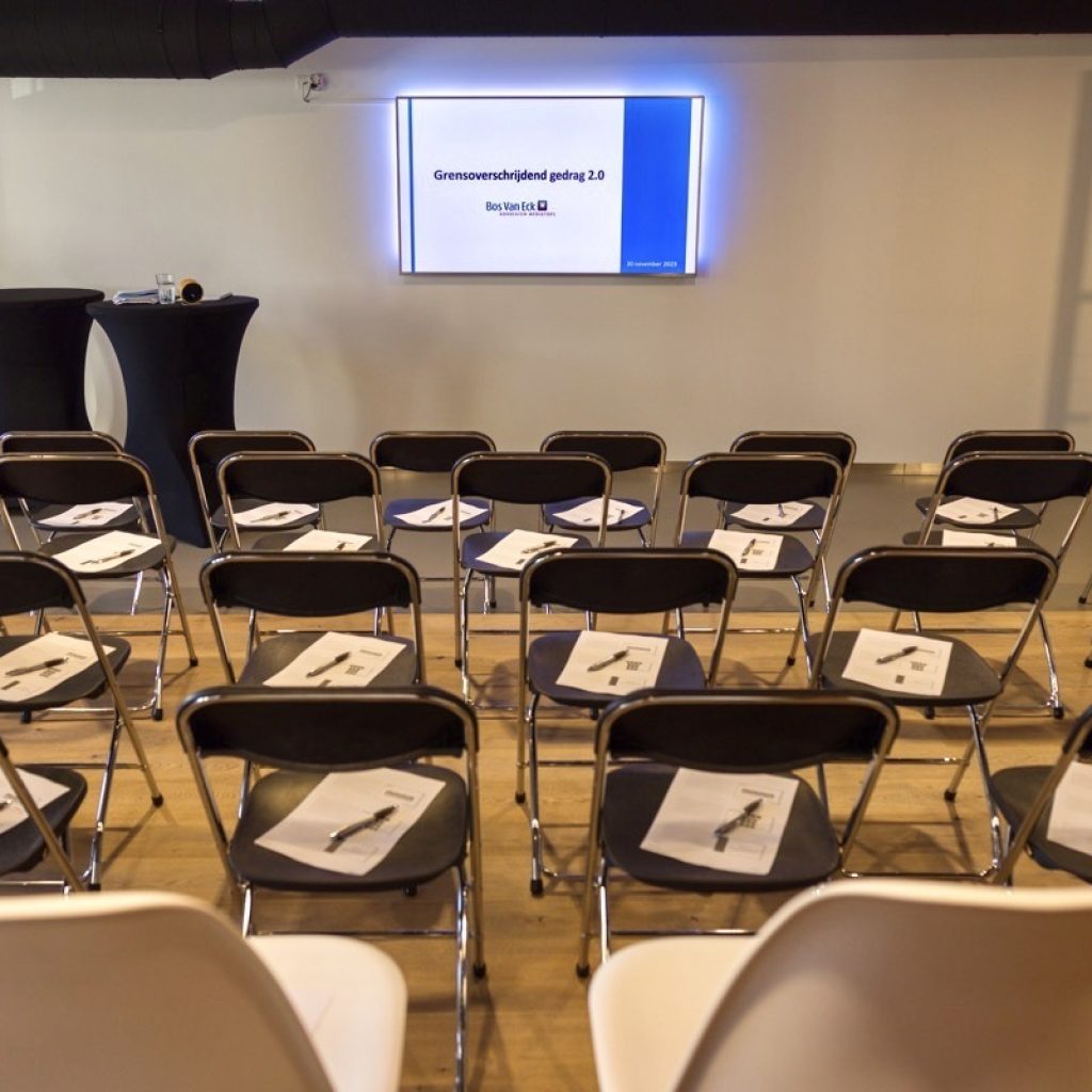 Conferentieruimte ingericht met stoelen en documenten die wachten op deelnemers voor een presentatie over Grensoverschrijdend gedrag 2.0 kennissessie op het scherm.