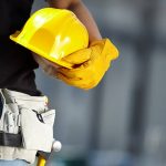 Een bouwvakker houdt een gele veiligheidshelm vast en draagt een gereedschapsriem, klaar voor de klus, ondanks de recente beëindiging van hun aannemingsovereenkomst.